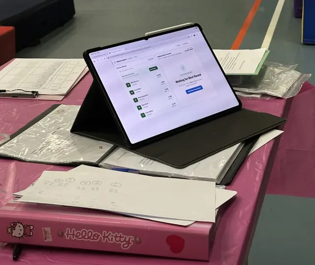 The Live Gym Scorer judge console on a tablet standing on a competition table beside paper score sheets
